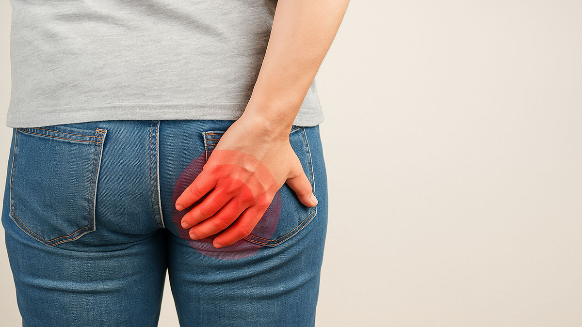 Rear view of a person in jeans holding their lower back or buttock with visible redness, symbolizing discomfort or pain from hemorrhoids.